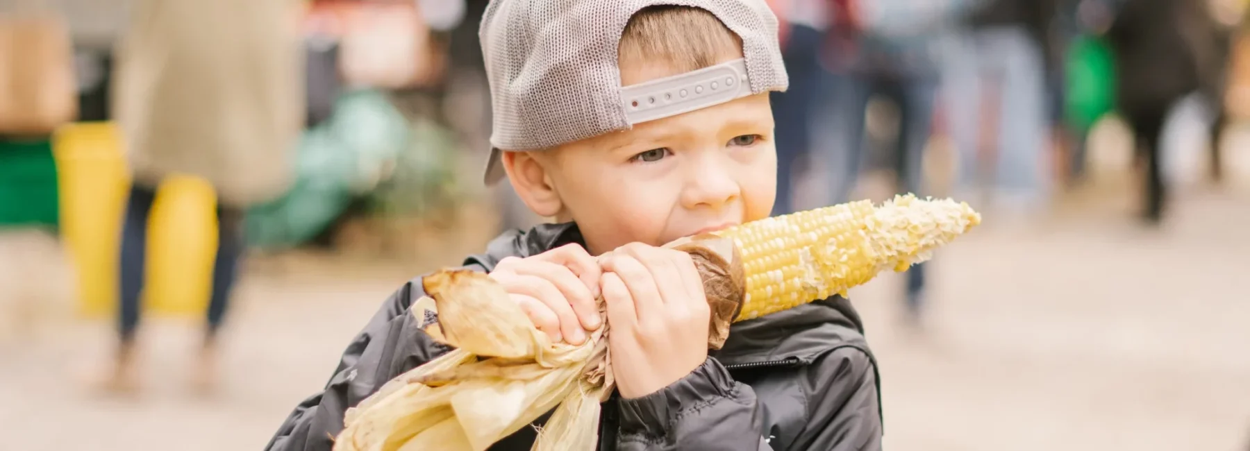 A child enjoying corn on the cob outdoors at a festival, surrounded by the vibrant culture and heritage of the region
